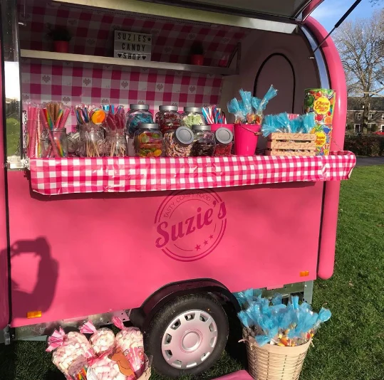 Remorque rose à l'enseigne Suzie's Candy Shop sur une pelouse, comptoir nappé de vichy couvert de bocaux de bonbons