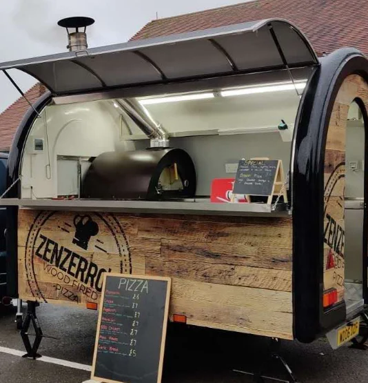 Remorque à bardage bois à l'enseigne Zenzerro, four à pizza à bois visible à l'intérieur et ardoise des prix posée devant